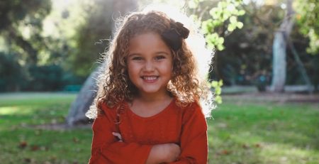 Niña pequeña con rizos rubios y vestido rojo, para de frente en plano medio corto. Con parque de fondo. Artículo Wola: La fuerza que te falta hoy. By Cely Cruz